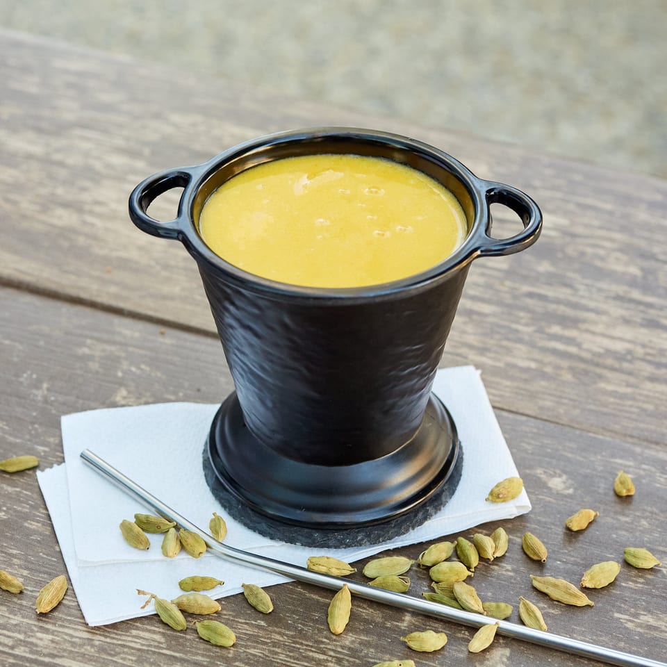 A black pot of yellow Indian curry, a metal straw, and cardamom pods on a wooden table.