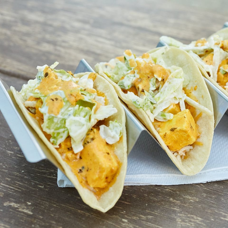 Three Indian-style tacos with paneer, slaw, and orange sauce in metal holders on a wooden table.