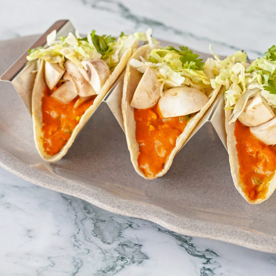 Three Indian-style tacos with orange sauce, mushrooms, and lettuce in a metal holder on a gray plate.