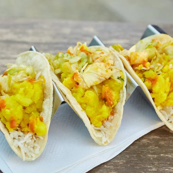 Three Indian-style tacos with yellow potato filling, rice, and cabbage, served in a metal holder.